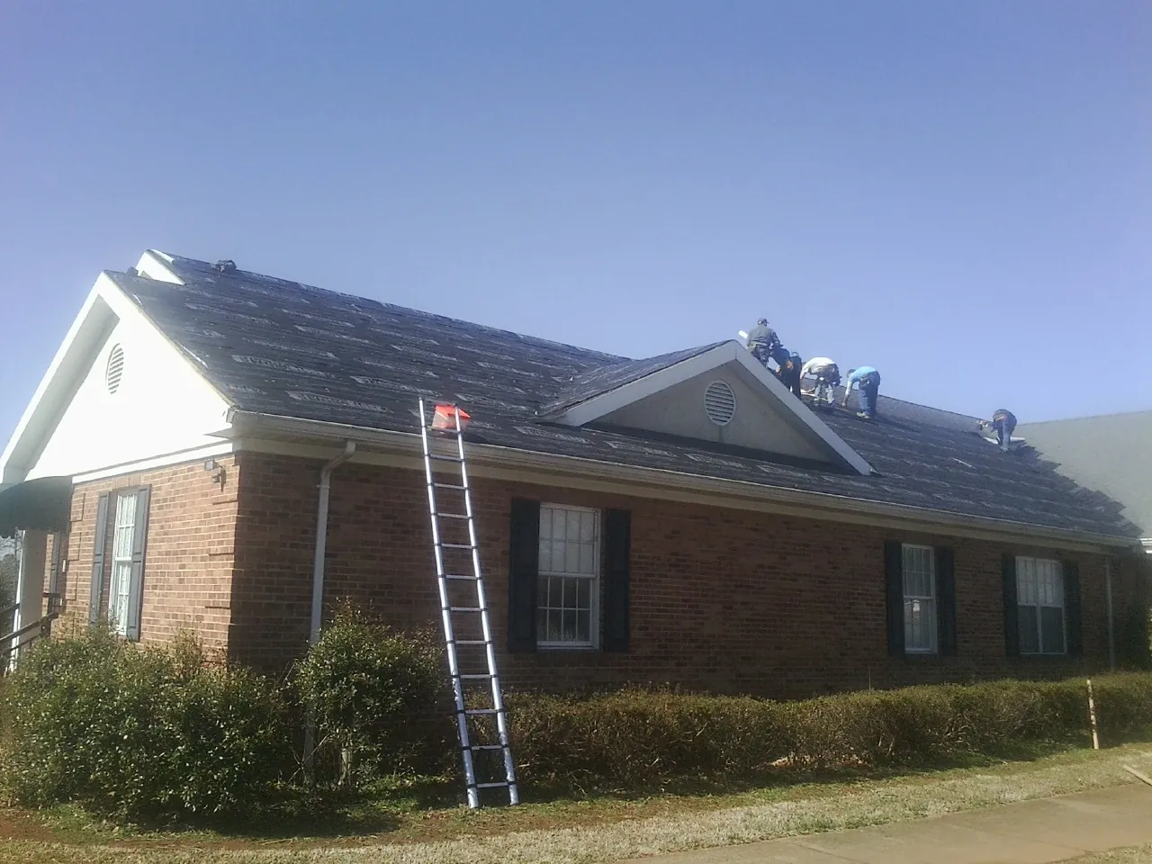 Crew tearing shingles off a brick ranch roof with a ladder set — Emergency Roof Repair work by our crew in West River