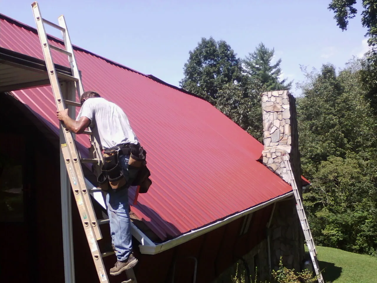 Roofer on a ladder beside a red metal roof and stone chimney — Soffit & Fascia Repair by Roofing Brothers in West River